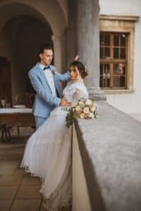 Ein Paar in formeller Kleidung posiert auf einem Steinbalkon und fängt die Essenz ihrer Hochzeit ein. Der Mann trägt einen hellblauen Anzug, während die Frau in ihrem weißen Spitzenhochzeitskleid einen Strauß pfirsichfarbener und weißer Blumen hält. Sie stehen inmitten historischer Architektur mit gewölbten Details und Holzfenstern.