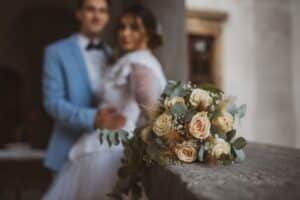Ein verschwommenes Paar steht dicht beieinander, mit einem Strauß aus beigen Rosen, Eukalyptus und kleinen weißen Blumen im Vordergrund – perfekt für ihre Hochzeit. Der Mann trägt einen hellblauen Anzug und die Frau ein weißes Kleid. Der Hintergrund ist sanft beleuchtet.
