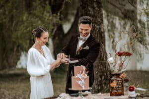 Das Paar strahlt, als es im Freien eine rosa Hochzeitstorte anschneidet und den Geist der Hochzeit verinnerlicht. Sie glänzt in einem weißen Kleid, während er in einem dunklen Anzug schick aussieht. Umgeben von Bäumen, dekorativen Blumen und durchscheinendem Stoff auf dem Tisch ist ihr Moment bildschön.