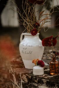 Eine große weiße Vase mit den Worten „To Have and To Hold“ enthält getrocknete Blumen und Gräser und verleiht eine romantische Note, die perfekt zu jeder Hochzeitsplanung passt. Sie steht auf einer verwitterten Steinoberfläche neben einer kleineren Vase mit orangefarbenen und roten Blumen und schafft so eine herbstliche Szene.