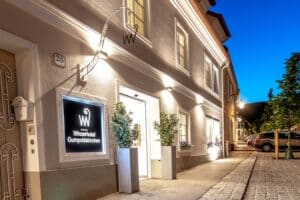 Nachtansicht eines gut beleuchteten Hoteleingangs mit einem Schild mit der Aufschrift „Winzerhotel Gumpoldskirchen“. Das Gebäude ist ideal zum Heiraten. Es verfügt über eine saubere, weiße Fassade mit Pflanzen am Eingang und einer Kopfsteinpflasterstraße davor. Perfekt für Ihre bezaubernde Hochzeitsplanung.