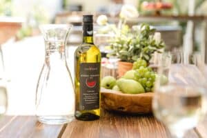 Eine Flasche Weißwein steht elegant auf einem Holztisch, perfekt für eine Hochzeitsfeier. Daneben glitzert eine Glaskaraffe mit Wasser im Licht. Daneben steht eine Holzschale, randvoll mit grünen Äpfeln und Trauben. Der sanft verschwommene Hintergrund aus Blumen sorgt für eine bezaubernde Hochzeitsatmosphäre.