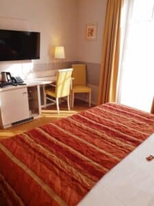 Ein gemütliches Hotelzimmer, perfekt für alle, die eine Hochzeit planen, mit einem ordentlich gemachten Bett mit einer orange-golden gemusterten Decke. An der Wand steht ein Schreibtisch mit den wichtigsten Annehmlichkeiten wie einer Lampe, einem Telefon und einem Wasserkocher. Darüber ist ein Flachbildfernseher angebracht, während Sonnenlicht durch das Fenster strömt.