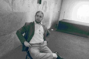Ein Graustufenbild zeigt einen nachdenklichen Mann, der auf einem Stuhl in einer Ecke des Raums sitzt und vielleicht über seine bevorstehende Hochzeit nachdenkt. Mit Bart und offenem Anzug scheint er vor den strukturierten Wänden in Gedanken versunken zu sein, als würde er über die Einzelheiten seiner Hochzeitsplanung nachdenken.