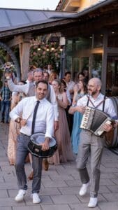 Eine fröhliche Hochzeitsfeier entfaltet sich mit einer Gruppe tanzender Menschen. Ein Mann spielt eine Trommel, während ein anderer vorne ein Akkordeon spielt. Die Gäste klatschen und lächeln, während ein Mann einen Blumenstrauß hochhält und die festliche Prozession im Freien anführt.