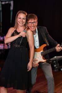 Eine Frau in einem schwarzen Kleid hält ein Mikrofon und lächelt, während sie neben einem Mann im Anzug steht, der E-Gitarre spielt. Sie treten auf einer Bühne mit dunklem Hintergrund auf und verleihen der Hochzeitsfeier des Paares eine magische Note.
