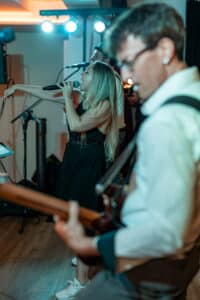 Eine Frau mit langen blonden Haaren singt leidenschaftlich in ein Mikrofon, während ein Mann im weißen Hemd im Vordergrund Gitarre spielt und die Bühne für eine unvergessliche Hochzeit bereitet. Der Raum ist schwach mit blauem Licht beleuchtet und schafft eine lebendige Atmosphäre, die perfekt ist, um die Liebe zu feiern.