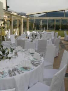 Eleganter Bankettsaal für eine Hochzeit. Jeder Tisch ist mit weißer Tischwäsche, ordentlich gefalteten Servietten, Kerzen und Glaswaren geschmückt. Große Fenster bieten einen malerischen Blick auf die Berge und das Grün draußen, perfekt für Ihre Hochzeitsplanungsträume.