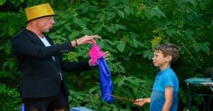 Ein Zauberer mit gelbem Hut führt einem Jungen mit einem Zauberstab in der Hand einen Zaubertrick mit rosa und blauen Tüchern vor. Ihr zauberhaftes Spektakel findet im Freien inmitten üppiger grüner Blätter statt und verleiht der Veranstaltung einen Hauch von Verspieltheit – perfekt für alle, die nach einzigartigen Ideen für die Hochzeitsplanung suchen.