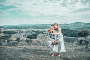 Braut und Bräutigam posieren glücklich im Freien, wobei die Braut den Bräutigam von hinten umarmt. Sie sitzen auf einem grasbewachsenen Hügel und verkörpern ihre Traumhochzeit, während sie unter einem teilweise bewölkten Himmel auf eine malerische Landschaft mit sanften Hügeln und Feldern blicken.