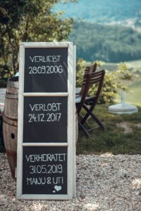Auf einem hübschen Kreidetafelschild im Freien sind wichtige Daten auf Deutsch zu sehen: „Verliebt 28.09.2006“, „Verlobt 24.12.2017“ und „Verheiratet 31.05.2019“ für Manu und Uti. Ein Herz symbolisiert ihre Liebesreise hin zu einer wunderschönen Hochzeit zwischen Holzstühlen und einer üppigen Landschaft im Hintergrund.
