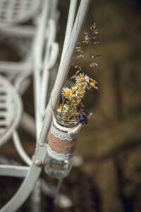 Ein kleiner Strauß Wildblumen in einem Glasgefäß hängt an einem weißen Metallstuhl, perfekt für eine Hochzeit. Die Blumen umfassen Gänseblümchen und andere zarte Blüten, und das Glas ist in Sackleinen und Spitze eingewickelt, was eine rustikale und charmante Dekoration ergibt, die ideal für Ihre Hochzeitsplanung ist.