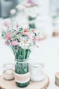 Ein rustikales Tischgesteck für eine Hochzeit besteht aus Wildblumen in einem Glasgefäß, das mit Schnur und Spitze umwickelt und auf einer Holzscheibe platziert ist. Der weiche Fokus im Hintergrund lässt weitere ähnliche Arrangements erkennen und schafft so ein charmantes, natürliches Ambiente, das perfekt für Ihre Hochzeitsplanung ist.