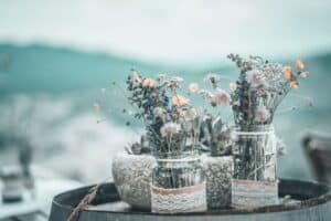 Zwei mit verschiedenen Wildblumen gefüllte, in Spitze eingewickelte Glasgefäße stehen auf einem Holzfass im Freien, perfekt für jede Hochzeit. Dahinter sorgen verschwommenes Grün und eine Hügellandschaft für eine ruhige Kulisse.