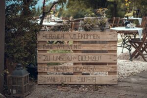 Ein Holzschild mit mehrsprachigem Text begrüßt die Gäste zur Hochzeit von „Manu + Uti“ am 31.05.19. Im Hintergrund sind Tische mit weißen Tischdecken elegant in Gartenatmosphäre gedeckt und zeugen von exquisiter Hochzeitsplanung.
