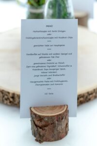 Eine rustikale Menükarte für eine Hochzeit, die auf einem Holzständer präsentiert wird, bietet eine Auswahl an Gerichten, darunter Suppen, Fisch mit Risotto und ein Fleischtrio mit verschiedenen Beilagen. Das Menü ist perfekt für Ihre Hochzeitsfeier und elegant in deutscher Sprache gestaltet.