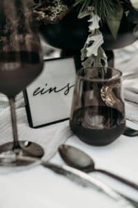 Eine Nahaufnahme eines gedeckten Tisches für eine Hochzeit zeigt ein Glas Rotwein, einen kleinen schwarzen Rahmen mit dem Wort „eins“ in Schreibschrift und Besteck auf einer weißen Tischdecke. Grünpflanzen zieren den Hintergrund und verleihen Ihrer perfekten Hochzeitsfeier Eleganz.