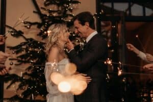 Ein Paar steht lächelnd vor einem geschmückten Weihnachtsbaum und stellt sich seine zukünftige Hochzeit vor, umgeben von Menschen mit Wunderkerzen. Die warme Umgebungsbeleuchtung schafft eine festliche Atmosphäre, die perfekt für die Planung ihrer Traumhochzeit ist.