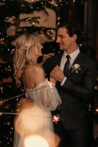 Ein Paar in Hochzeitskleidung steht vor einem geschmückten Weihnachtsbaum und verkörpert die Freude des Heiratens. Sie blicken sich liebevoll an, umgeben von warmen Lichtern und festlichen Dekorationen, die die gemütliche, romantische Atmosphäre verstärken – ein bildschöner Moment in ihrer Hochzeitsplanung.