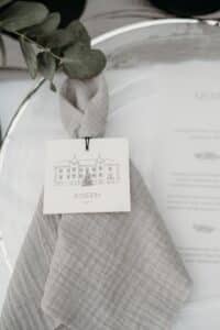 Eine hellgraue Serviette, elegant gefaltet und mit einer Karte mit der Aufschrift „Joseph“ zusammengebunden, zeigt eine detaillierte Gebäudeskizze. Perfekt für eine Hochzeit, liegt die Serviette auf einem weißen Teller mit einem Menü, das durch Grünpflanzen obenauf noch aufgewertet wird.