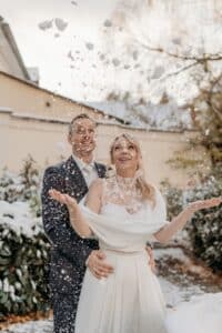 Ein Paar in formeller Kleidung steht draußen, umgeben von schneebedeckten Pflanzen, und verkörpert die Romantik einer Winterhochzeit. Die Frau wirft freudig Schnee in die Luft, während der Mann sie von hinten umarmt, beide lächeln. Die Szene fängt die Essenz einer hellen und winterlichen Liebesfeier ein.