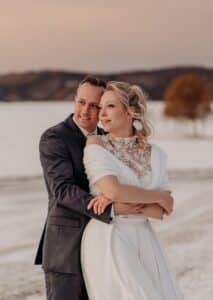 Ein Paar steht bei Sonnenuntergang in einer verschneiten Landschaft, eingehüllt in die bezaubernde Aura einer Hochzeit. Die Frau, in einem weißen Kleid mit Blumendetails und einem Schal, wird von dem Mann im dunklen Anzug umarmt. Mit heiterem Gesichtsausdruck blicken sie in die Ferne und stellen sich ihre Reise nach der Hochzeit vor.