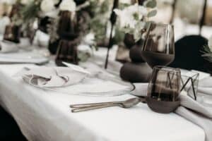 Elegante Tischdekoration mit dunklem Glasgeschirr, weißen Tellern und gefalteten Servietten auf einer weißen Tischdecke. Der Tisch ist perfekt für eine Hochzeit, er ist mit Blumenarrangements und Grünpflanzen geschmückt und schafft eine anspruchsvolle und stilvolle Atmosphäre, die ideal für die Hochzeitsplanung ist.