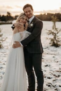 Ein Paar im Hochzeitskleid umarmt sich zärtlich in einer verschneiten Landschaft und verkörpert die Essenz einer Hochzeit. Die Braut trägt ein weißes Kleid und der Bräutigam einen dunklen Anzug. Bäume und ein bewölkter Himmel bilden den Hintergrund und schaffen eine ruhige Winterlandschaft, die perfekt für diejenigen ist, die eine durchdachte Hochzeitsplanung in Betracht ziehen.