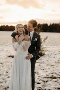 Ein Paar steht bei Sonnenuntergang auf einem verschneiten Feld, eingefangen in einem perfekten Hochzeitsmoment. Die Frau im weißen Kleid lacht, als der Mann im Anzug sie von hinten umarmt. Der Himmel ist in Orange- und Rosatönen erleuchtet und Bäume säumen den Horizont.