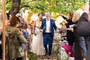 Ein fröhliches Brautpaar schreitet im Freien, umgeben von Gästen, den Gang entlang. Als Teil ihrer Hochzeit trägt die Braut ein weißes Kleid und der Bräutigam einen blauen Anzug. Seifenblasen schweben in der Luft und die Szene ist voller Lächeln und Feierlichkeiten.