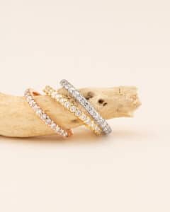 Vier funkelnde Ringe, perfekt für eine Hochzeit, werden elegant auf Treibholz präsentiert. Die Ringe sind mit kleinen Diamanten verziert und variieren in den Farben Roségold, Gelbgold und Silber. Ihr Glanz bildet einen auffälligen Kontrast zum hellbeigen Hintergrund und bereitet die Bühne für eine unvergessliche Feier.