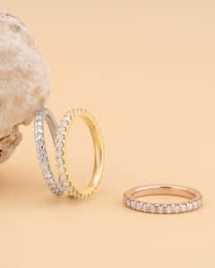 Drei mit Diamanten besetzte Ringe, perfekt für die Hochzeitsplanung, werden vor einem beigen Hintergrund präsentiert. Die Ringe in Silber, Gold und Roségold weisen jeweils eine durchgehende Reihe kleiner funkelnder Diamanten auf. Links ist ein strukturierter Stein positioniert.