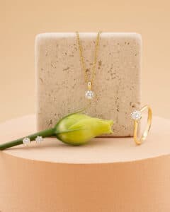 Eine Schmuckpräsentation, die sich perfekt für eine Hochzeit eignet, besteht aus einer goldenen Halskette, Ohrringen und einem Ring, die jeweils mit einem funkelnden Diamanten besetzt sind. Eine gelbe Rosenknospe ruht neben dem Schmuck auf einem runden Sockel vor einem beigen Hintergrund.