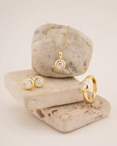 Ein atemberaubendes Goldschmuckset, perfekt für eine Hochzeit, exquisit präsentiert auf beigen Steinen. Es umfasst eine Halskette mit einem runden Diamantanhänger, Diamantohrstecker und einen Diamantring. Das glänzende Gold bildet einen eleganten Kontrast zur rauen Textur der Steine.