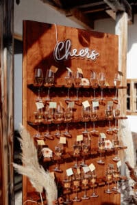 Eine hölzerne Schautafel mit mehreren Ablagen hält Weingläser, die mit einem leichten Getränk gefüllt sind, perfekt für jede Hochzeit. An jedem Glas ist eine kleine gelbe Karte befestigt. Oben steht auf einem beleuchteten Schild „Prost“. Pampasgras schmückt die Szene und verleiht Ihrer Hochzeitsplanung Eleganz.