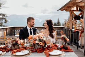 Ein Paar in Anzug und Brautkleid sitzt an einem wunderschön dekorierten Tisch im Freien und verkörpert damit die Essenz perfekter Hochzeitsplanung. Der Tisch ist mit farbenfrohen Blumenarrangements und elegantem Geschirr geschmückt, und die malerische Berglandschaft bildet die Kulisse ihrer Hochzeit.