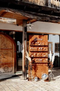 Eine rustikale Hochzeitsdekoration vor einem Veranstaltungsort besteht aus einem Holzbrett mit Champagnergläsern und einem „Prost“-Schild. Pampasgras schmückt das Ganze und reflektierende Diskokugeln glitzern am Sockel neben einer großen Holztür. Die perfekte Inspiration für alle, die sich intensiv mit der Hochzeitsplanung beschäftigen.