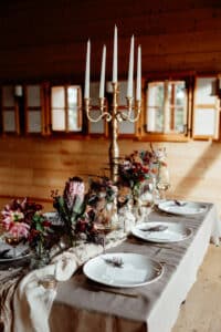 Ein rustikaler Esstisch ist für eine Hochzeit gedeckt, mit eleganten weißen Tellern, Blumenarrangements und einem hohen goldenen Kandelaber, der die Szene erhellt. Das gemütliche Holzinterieur mit mehreren Holzfenstern bietet die perfekte Kulisse für Heiratsfeiern.