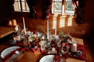 Ein warm beleuchteter Holztisch für ein gemütliches Abendessen erinnert an die Romantik einer Hochzeitsfeier. Kerzen, Blumen, silberne Ornamente und ein roter Tischläufer verleihen Eleganz, während sanftes Licht durch Fenster mit roten Vorhängen fällt und eine einladende Atmosphäre schafft, die perfekt für eine Hochzeit ist.
