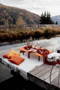 Eine gemütliche Außeneinrichtung auf einem Holzsteg über dem Wasser bietet die perfekte Kulisse für eine intime Hochzeitsplanung mit roten und orangefarbenen Kissen, einer weißen Decke und einem Tisch mit Snacks und Getränken. Herbstbäume und ein alter Holzzaun umgeben den Steg, im Hintergrund eingerahmt von majestätischen Bergen.