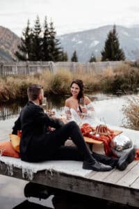 Ein Paar genießt ein malerisches Picknick an einem Teich und sitzt auf einem mit Kissen und Decken geschmückten Holzsteg. In formeller Kleidung schwelgen sie im Rahmen ihrer Hochzeitsfeier in der Ruhe der Berge und Bäume, wobei eine skurrile Diskokugel ihrem besonderen Tag Charme verleiht.