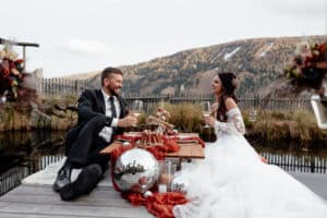 Braut und Bräutigam sitzen auf einem Holzsteg in der Nähe eines Teichs und genießen ein Picknick mit Wein und Dekoartikeln. Der Bräutigam trägt einen schwarzen Anzug, die Braut ein weißes Kleid. Berge und Laub vervollständigen die ruhige Kulisse, perfekt für eine intime Hochzeit als Teil ihrer durchdachten Hochzeitsplanung.