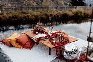 Eine gemütliche Tischdekoration im Freien an einem Teich, perfekt für eine intime Hochzeitsfeier, mit roten und orangefarbenen Kissen, einem Holztisch mit festlichen Desserts, Kerzen und zwei Weingläsern. Über dem Tisch hängt ein weißes Tuch, im Hintergrund sind Discokugeln und Herbstlaub zu sehen.