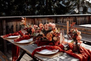 Ein rustikaler Esstisch im Freien ist elegant für eine Hochzeit gedeckt, mit Blumenarrangements in Rosa- und Orangetönen, umgeben von Herbstblättern. Rote Stoffläufer betonen den Holztisch, der mit Kerzen und ordentlich arrangiertem Geschirr geschmückt ist, perfekt für eine bezaubernde Hochzeitsfeier.