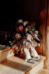Ein Strauß gemischter Blumen und ein Paar silberner High Heels stehen auf Holzstufen, perfekt für eine Hochzeit. Das Sonnenlicht wirft einen warmen Schein, der die Texturen und Farben der Blumen und den Glanz der Schuhe hervorhebt und eine bezaubernde Bühne für die Hochzeit bietet.