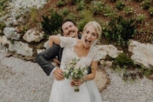 Ein glückliches Paar posiert im Freien bei seiner Hochzeit. Die Braut in einem weißen Spitzenkleid hält einen Blumenstrauß und lächelt breit, während der Bräutigam sie von hinten umarmt. Sie stehen auf einem Kiesweg, umgeben von Grün und Felsen, und verkörpern die Glückseligkeit des Heiratens.