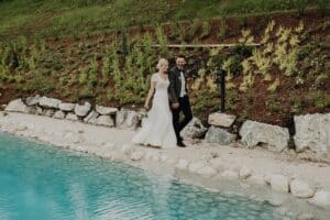 Eine Braut im weißen Kleid und ein Bräutigam im Anzug gehen Hand in Hand am Rand eines Pools entlang, der von Felsen und Grün umgeben ist. Die Szene ist ruhig und fängt die Essenz des Hochzeitsglücks ein, mit üppigen Pflanzen auf einem sanften Hang im Hintergrund.