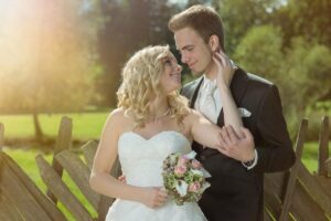 Braut und Bräutigam stehen eng beieinander und lächeln sich an ihrem Hochzeitstag an. Die Braut hält einen Strauß rosafarbener und weißer Blumen, während der Bräutigam einen dunklen Anzug trägt. Sie stehen neben einem Holzzaun mit einem grünen, sonnenbeschienenen Hintergrund, der perfekt ist, um ihre Hochzeitsplanungsvision festzuhalten.