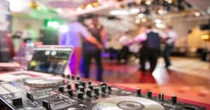 Ein DJ-Setup mit Plattenspieler und Laptop im Vordergrund schafft die Bühne für eine lebendige Hochzeitsplanungsszene. Im Hintergrund tanzen Gäste in einem farbenfrohen, sanft beleuchteten Raum und schaffen so eine lebendige und festliche Hochzeitsatmosphäre.