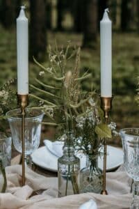 Eine rustikale Tischdekoration im Freien, perfekt für eine Hochzeit, mit hohen weißen Kerzen in goldenen Haltern, klaren Glastellern und zarten Blumenarrangements in Glasvasen. Der Hintergrund bietet einen wunderschön verschwommenen Blick auf einen Wald.